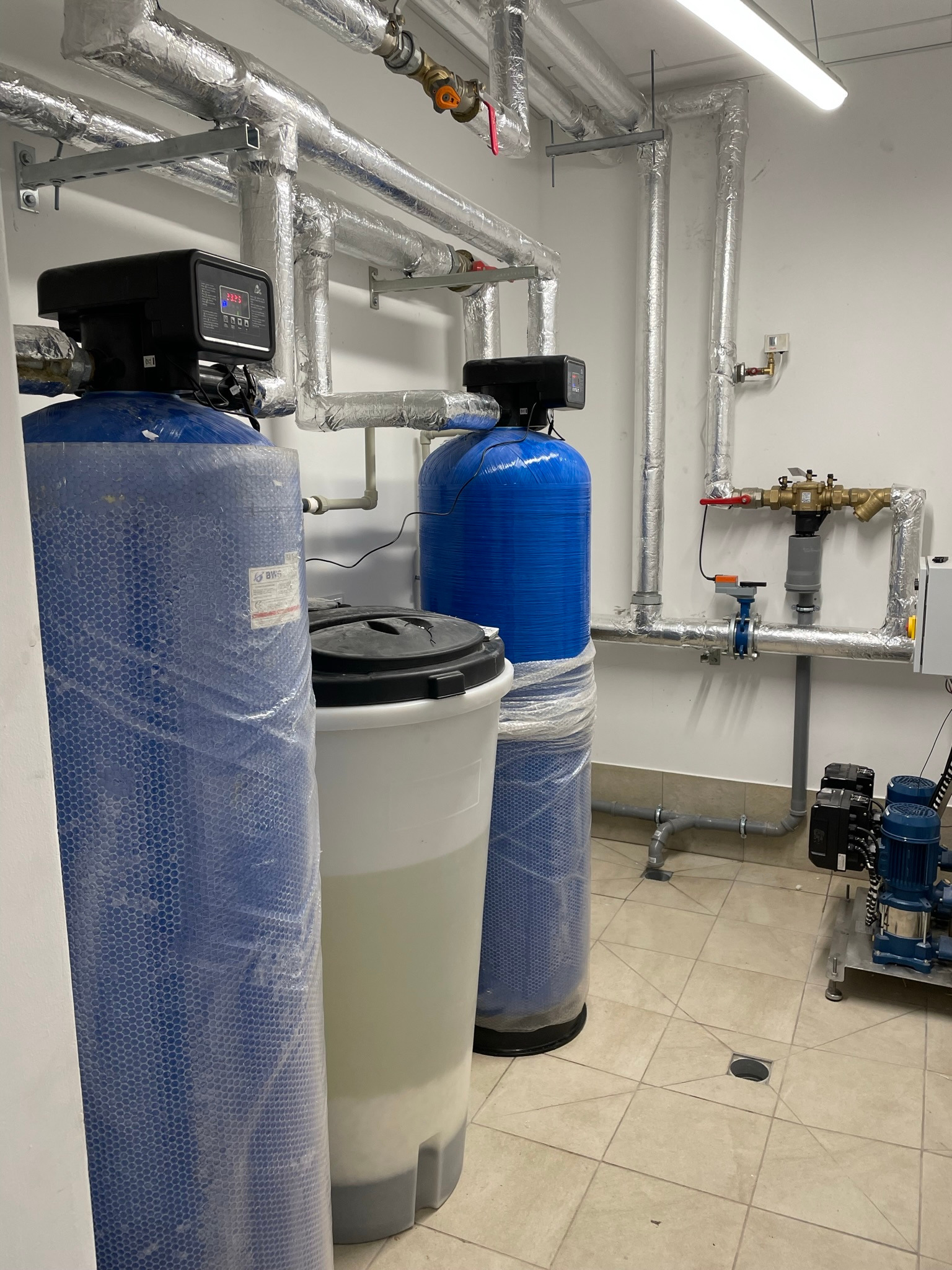 Dwa niebieskie filtry wody z czarnymi głowicami i wyświetlaczami, biały zbiornik z czarną pokrywą, rury w izolacji termicznej, instalacja wodna z zaworami, oraz pompy na płytce w pomieszczeniu...