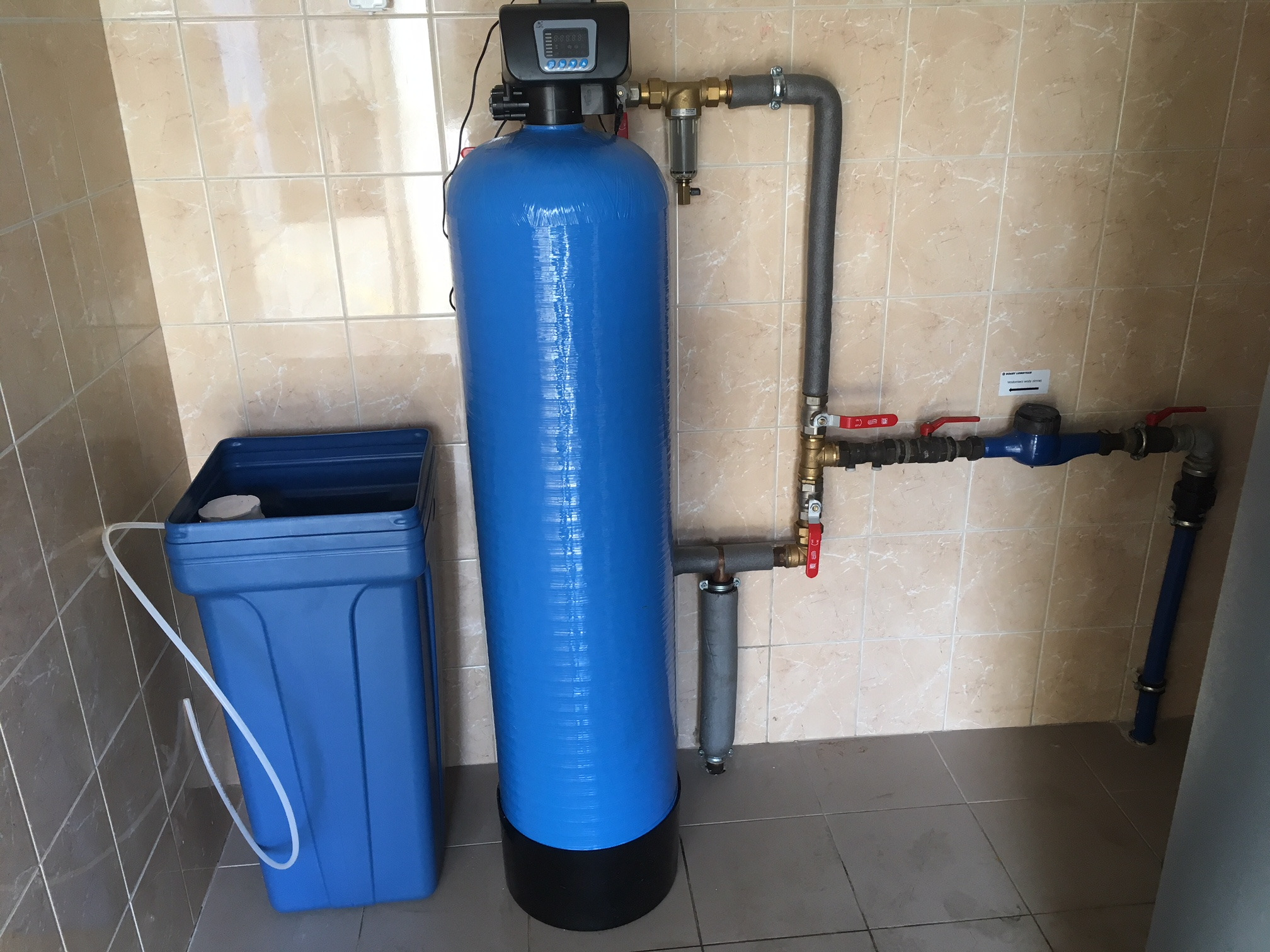 Niebieski zbiornik z elektronicznym sterownikiem i orurowaniem w pomieszczeniu wyłożonym płytkami, obok stoi niebieski pojemnik z rurką.