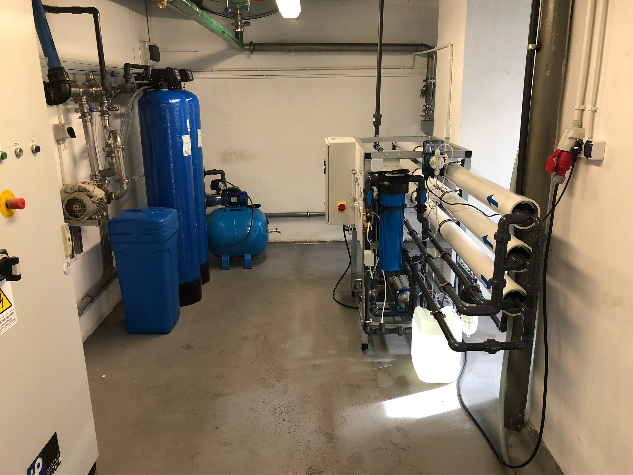 Kompleksowa instalacja przemysłowa z niebieskimi zbiornikami ciśnieniowymi, pompami i systemem filtrów membranowych w sterylnej hali, widoczne liczne rury i zawory.
