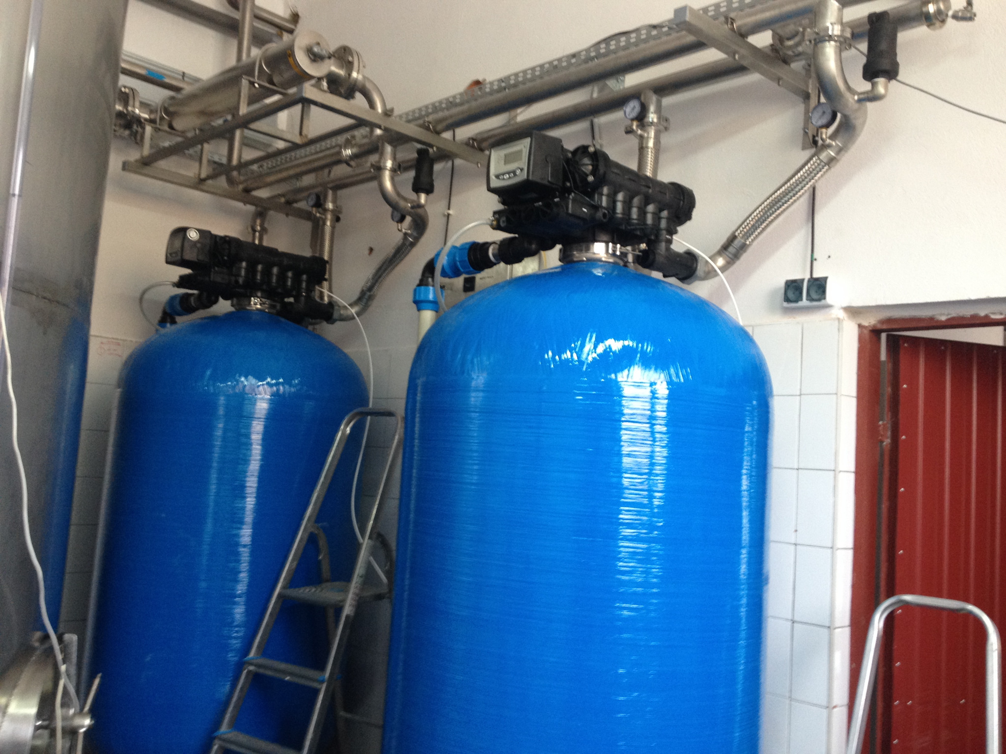 Dwa niebieskie zbiorniki ciśnieniowe z czarnymi głowicami sterującymi, połączone rurami ze stali nierdzewnej, ustawione w pomieszczeniu z białą glazurą i drabiną inspekcyjną.
