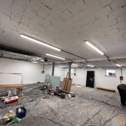 atcc z.o.o - Przestronne pomieszczenie w trakcie remontu: zabezpieczona folią podłoga, widoczne narzędzia, pędzle, drabina, płyty gipsowe, worki z zaprawą oraz mężczyzna używający szlifierki przy ścianie.
