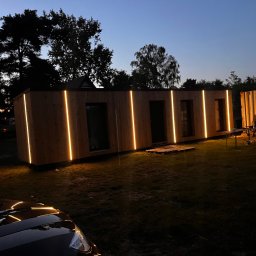 atcc z.o.o - Nowoczesny dom modułowy z drewnianą elewacją i pionowym oświetleniem LED, sfotografowany o zmierzchu na tle drzew i kontenera, z widocznym fragmentem samochodu w dolnej części kadru.