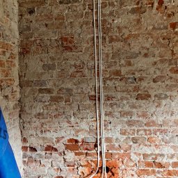 atcc z.o.o - Dwa białe kable elektryczne prowadzone pionowo wzdłuż ściany z czerwonej cegły w trakcie remontu, widoczny fragment niebieskiej folii ochronnej.