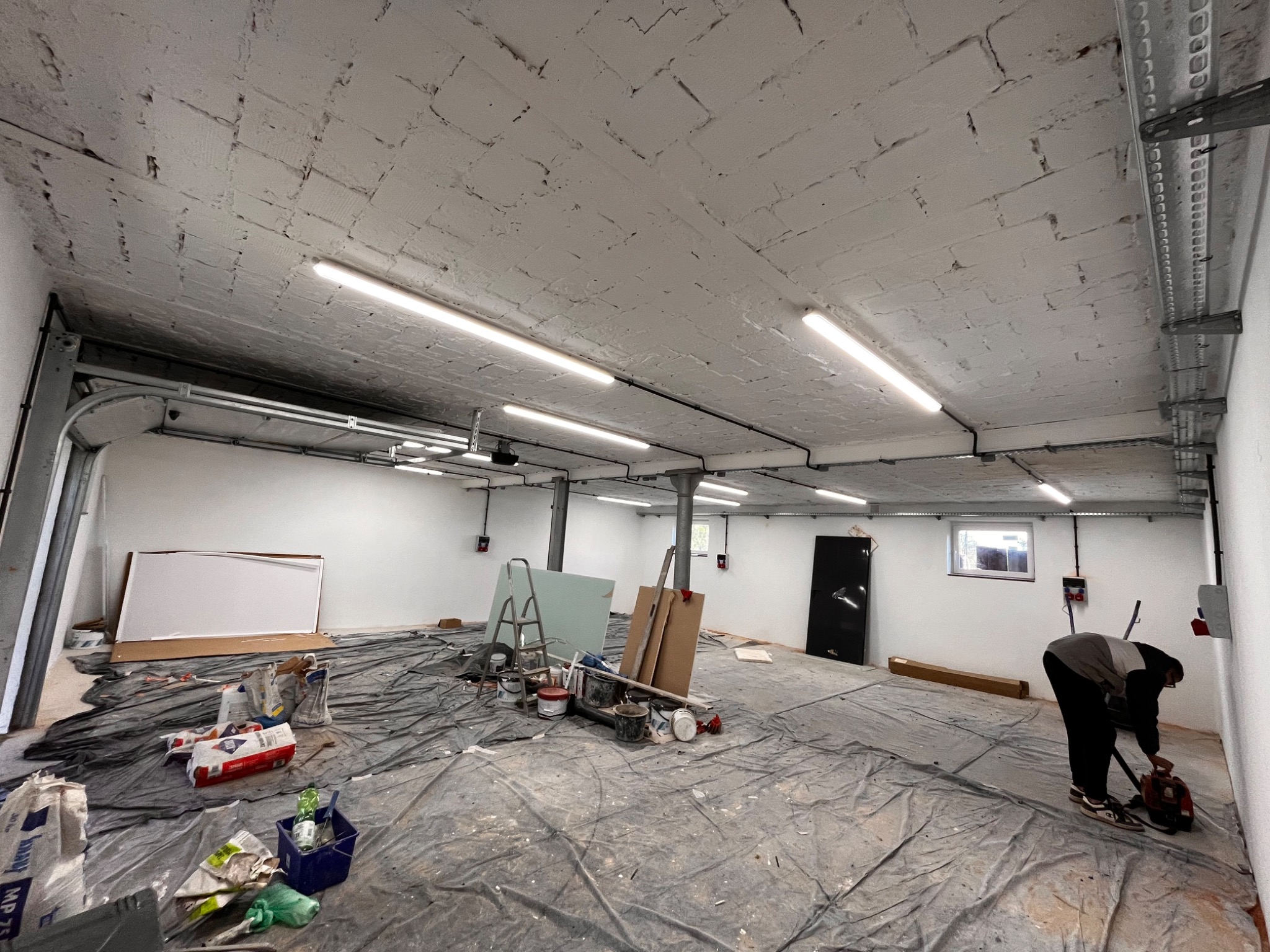 Przestronne pomieszczenie w trakcie remontu: zabezpieczona folią podłoga, widoczne narzędzia, pędzle, drabina, płyty gipsowe, worki z zaprawą oraz mężczyzna używający szlifierki przy ścianie.