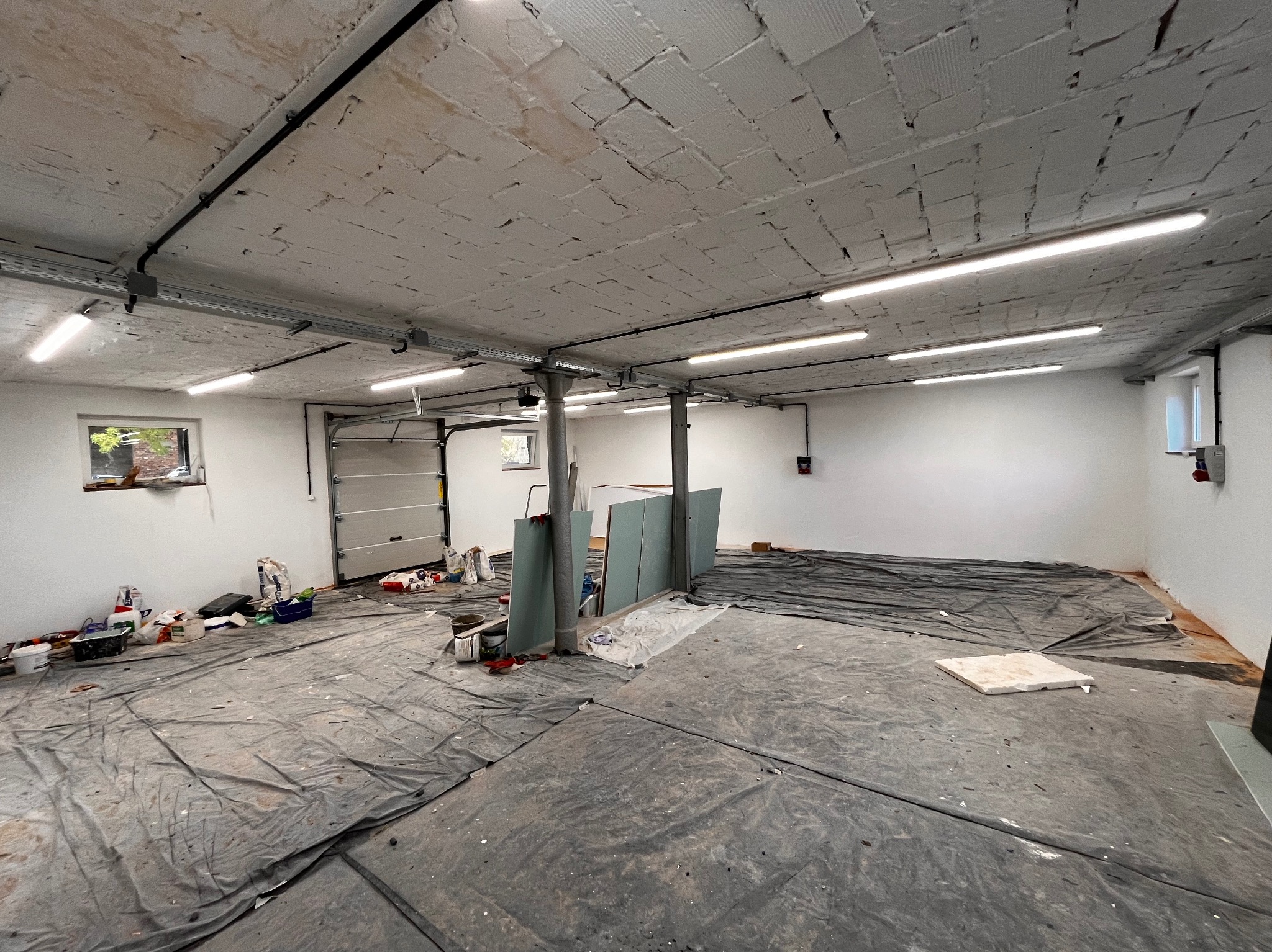 Przestronne pomieszczenie w trakcie remontu, zabezpieczone folią malarską, widoczne instalacje elektryczne pod sufitem, przygotowane płyty gipsowo-kartonowe i materiały malarskie.