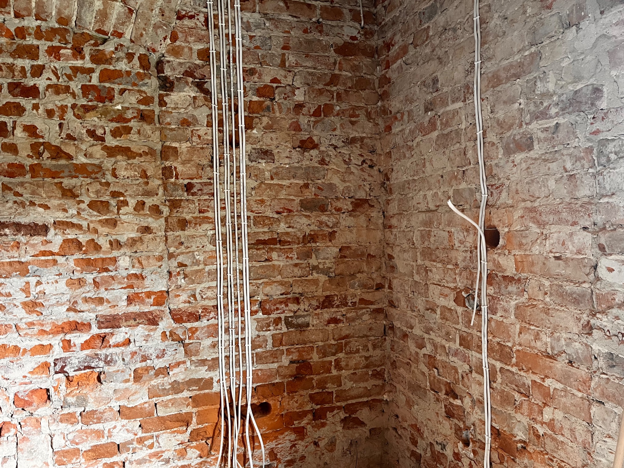 Surowe wnętrze z ceglanymi ścianami, na których widać pionowo poprowadzone białe kable elektryczne, mocowane do ściany za pomocą uchwytów. Fragment ściany z widoczną dziurą po instalacji.