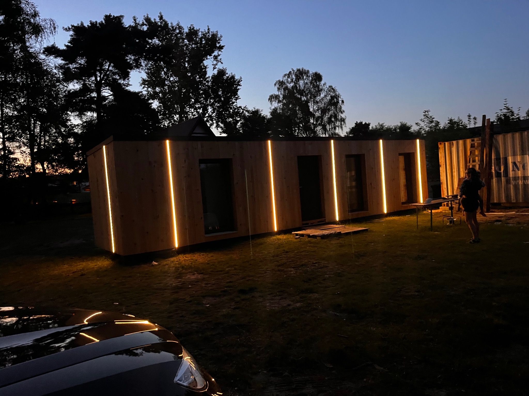 Nowoczesny dom modułowy z drewnianą elewacją i pionowym oświetleniem LED, sfotografowany o zmierzchu na tle drzew i kontenera, z widocznym fragmentem samochodu w dolnej części kadru.