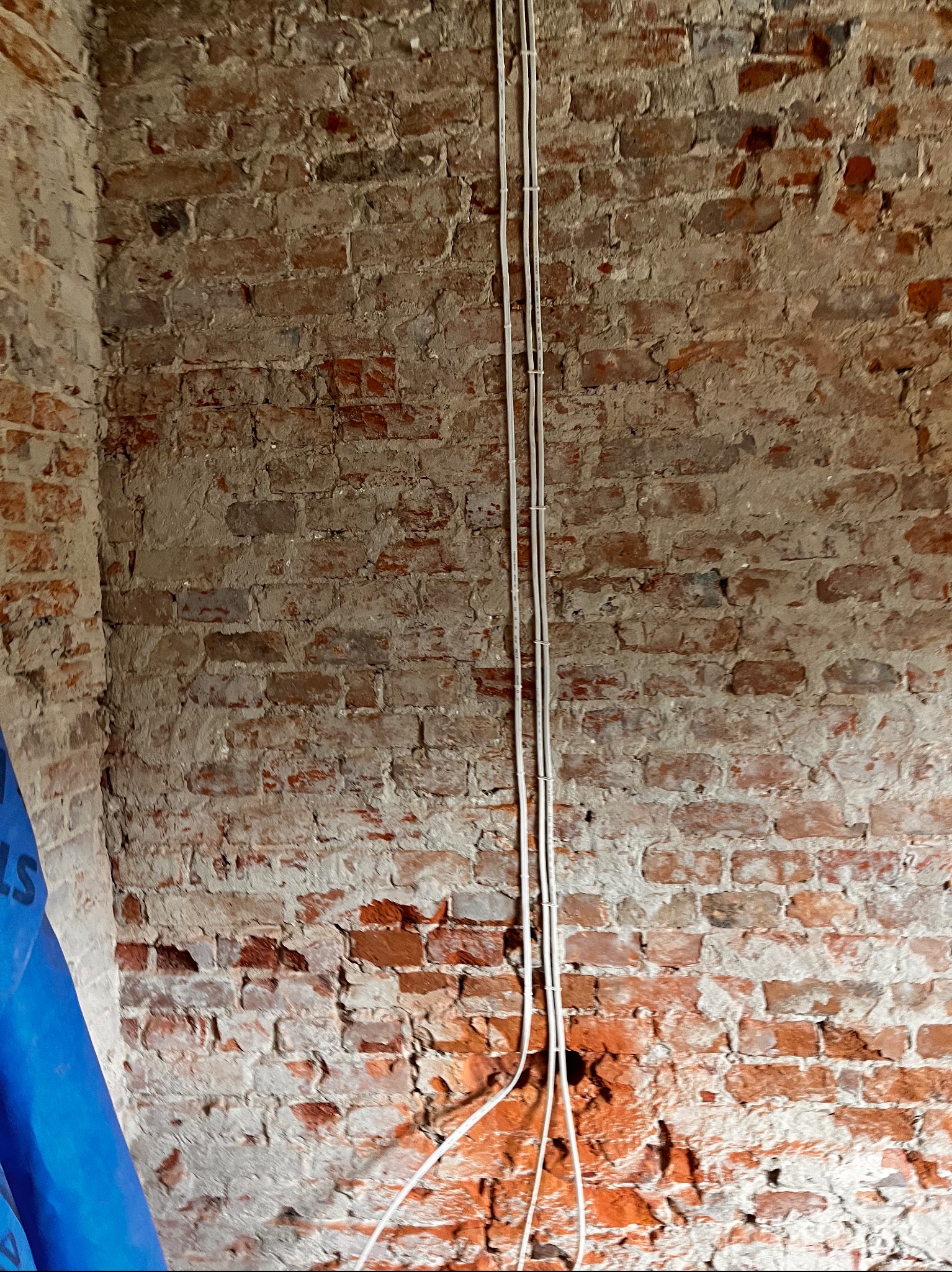 Dwa białe kable elektryczne prowadzone pionowo wzdłuż ściany z czerwonej cegły w trakcie remontu, widoczny fragment niebieskiej folii ochronnej.