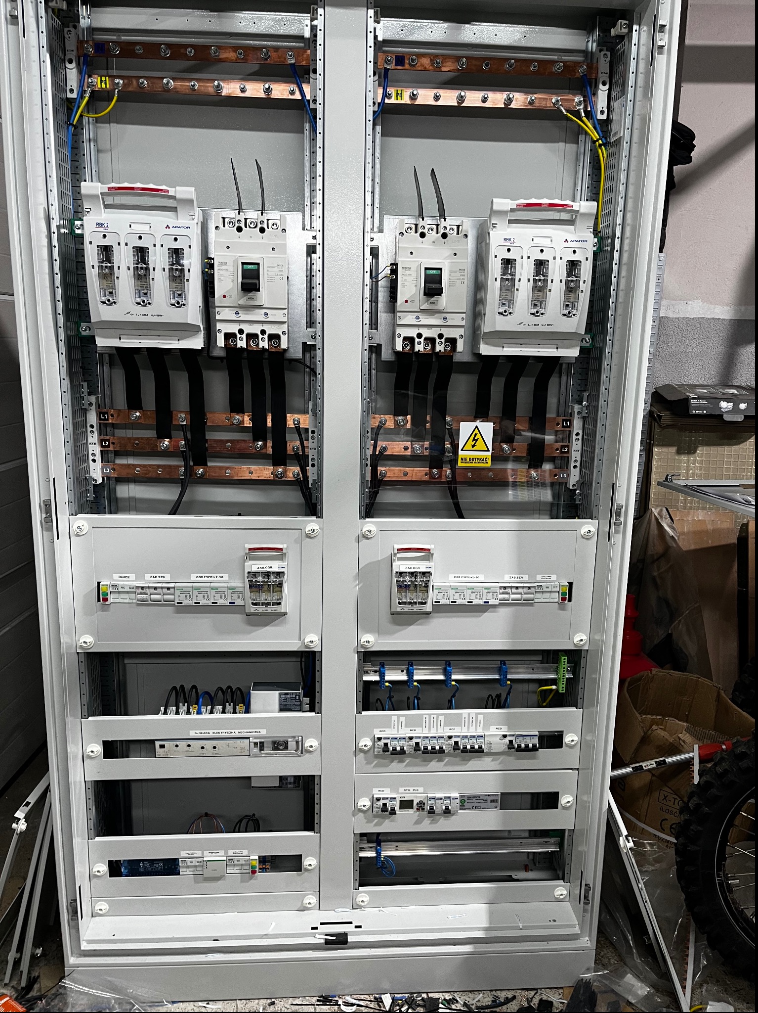 Otwarta szafa elektryczna z widocznymi bezpiecznikami, wyłącznikami, szynami miedzianymi i okablowaniem, z naklejką ostrzegawczą 'Nie dotykać!', obok widoczne narzędzia i koło motocyklowe.