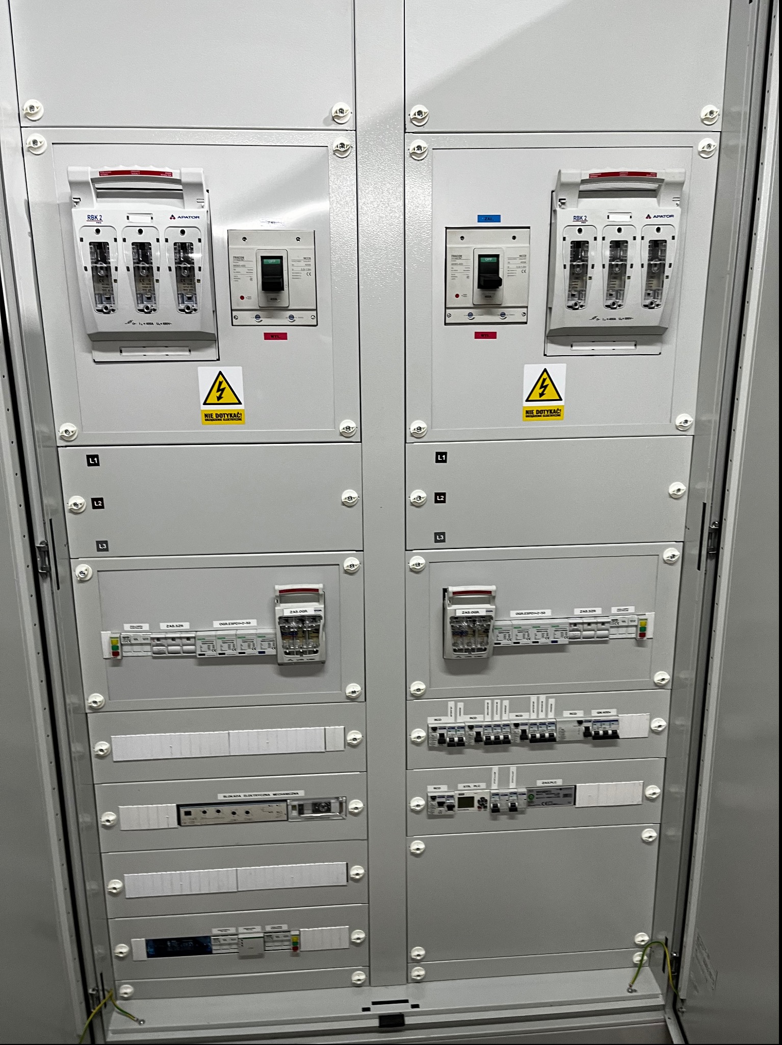 Szara szafa elektryczna z bezpiecznikami, wyłącznikami i oznaczeniami faz L1, L2, L3 oraz ostrzeżeniami 'NIE DOTYKAĆ!'