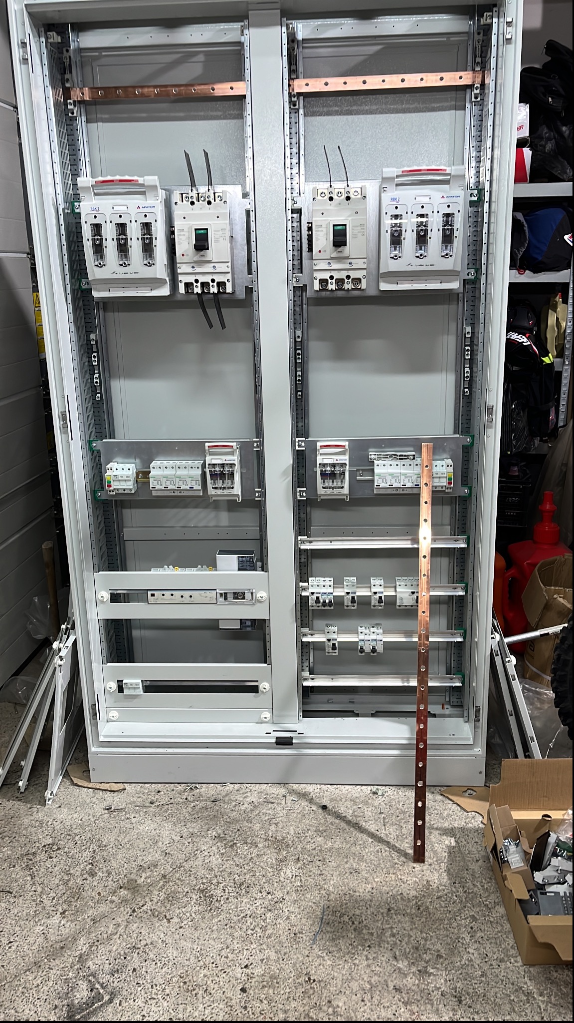 Rozdzielnia elektryczna w trakcie montażu, widoczne szyny miedziane, bezpieczniki i inne elementy instalacji elektrycznej w szarej obudowie. Na podłodze widoczny element miedziany i karton...