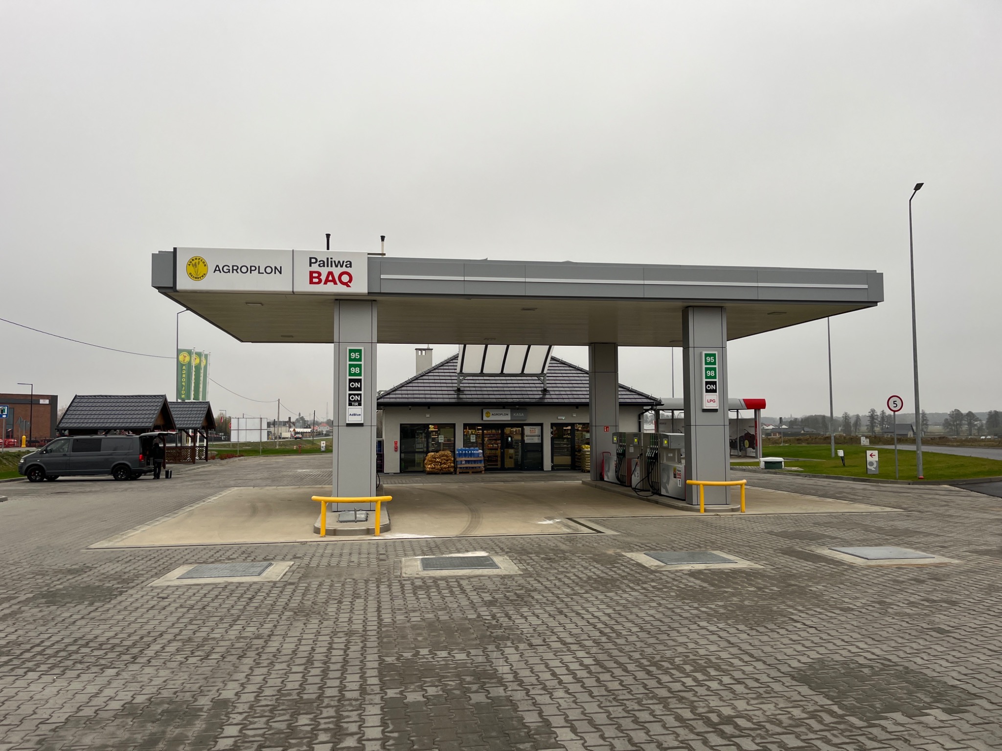 Stacja paliw Agropolon BAQ z szarym vanem zaparkowanym obok, widok na budynek stacji i dystrybutory paliwa na tle pochmurnego nieba.