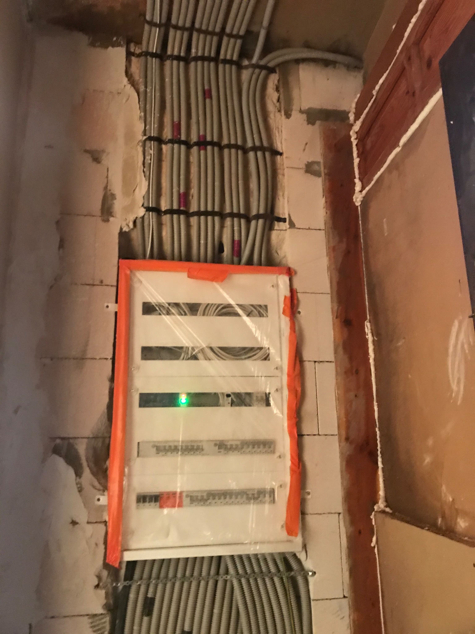 Rozdzielnica elektryczna zabezpieczona folią i pomarańczową taśmą, widoczne kable w peszlach mocowane do ściany, ściana z pustaków w trakcie prac wykończeniowych.
