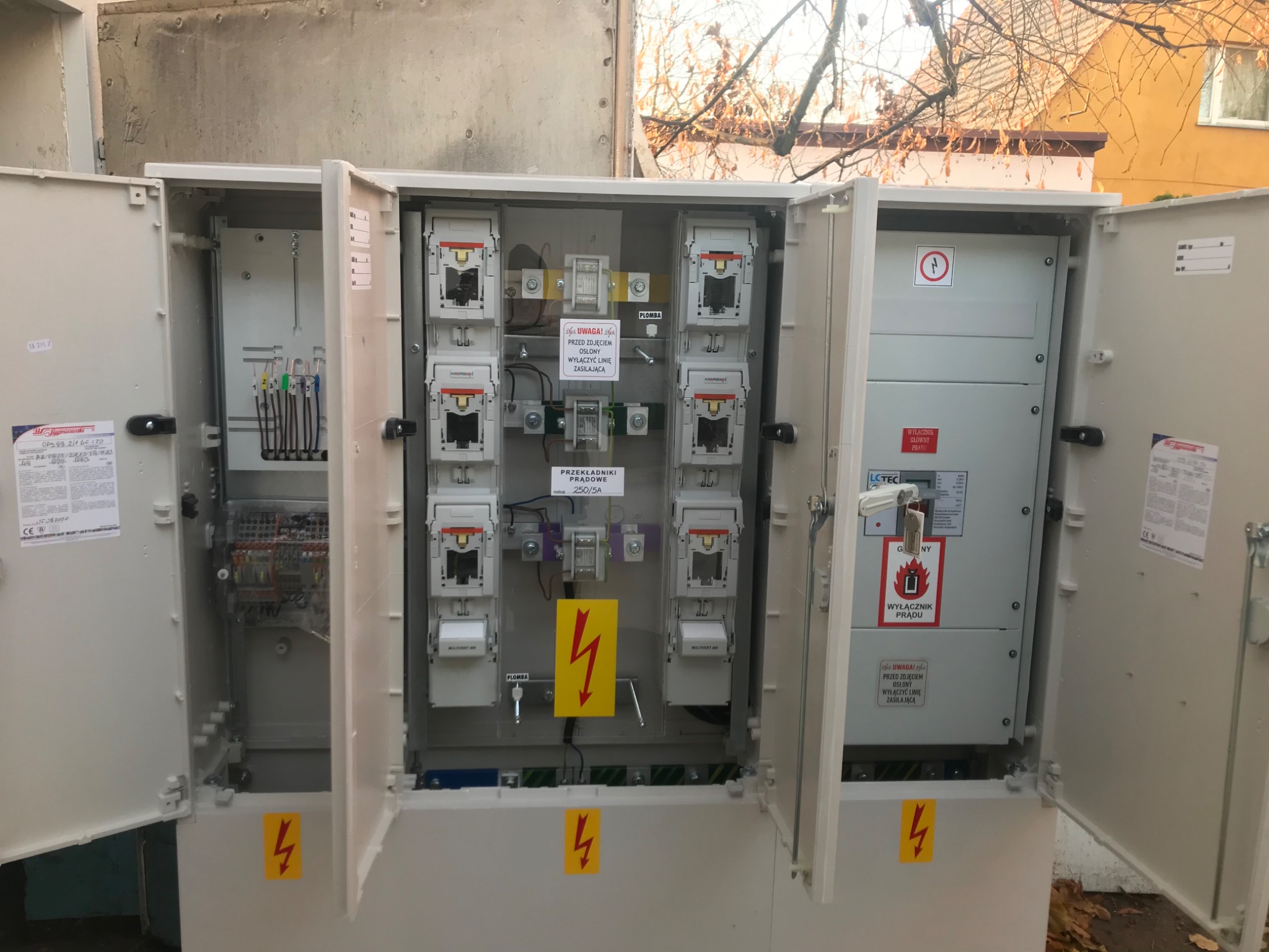 Otwarta szafa elektryczna na zewnątrz budynku, widoczne zabezpieczenia, przekładniki prądowe, wyłączniki, okablowanie i naklejki ostrzegawcze z symbolem błyskawicy.