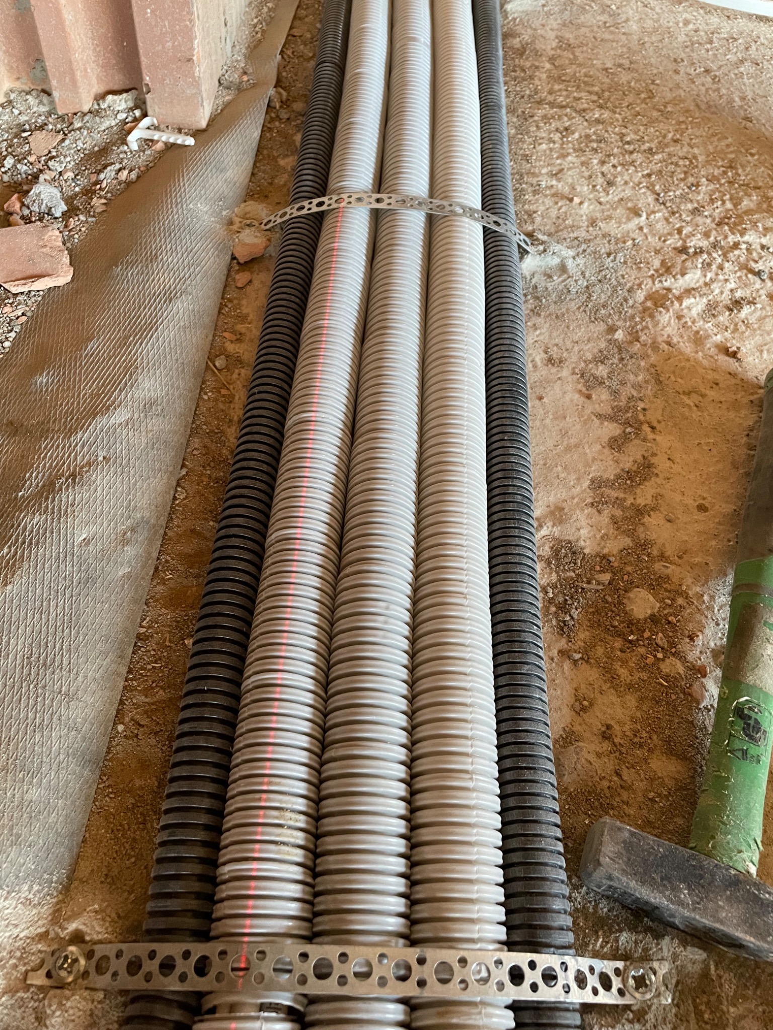 Ułożone w wykopie rury karbowane: trzy szare i dwie czarne, spięte metalową listwą perforowaną, obok fragment muru z cegły i zielony młotek.