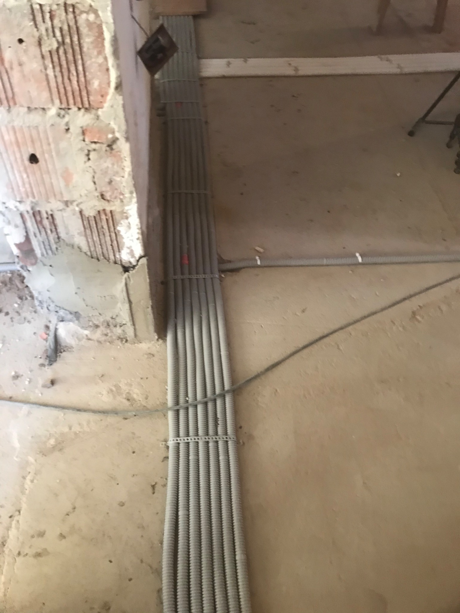 Wiązka szarych, karbowanych rur instalacyjnych przymocowana do ściany z surowej cegły, widoczne elementy konstrukcyjne i zaprawa, podłoga betonowa w tle, etap prac wykończeniowych.