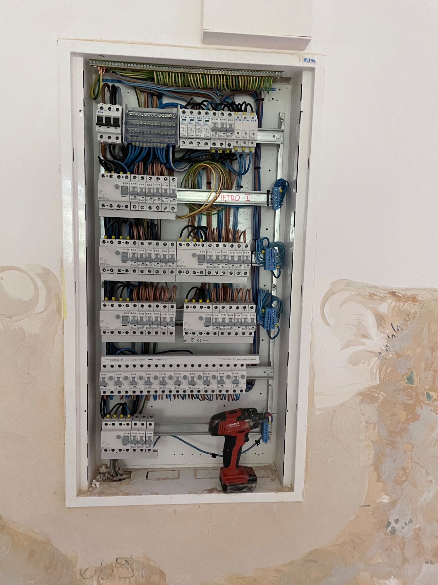 Otwarta skrzynka z bezpiecznikami i okablowaniem w trakcie montażu, widoczna czerwona wiertarko-wkrętarka Hilti na dole, ściana w tle z widocznymi śladami po remoncie.