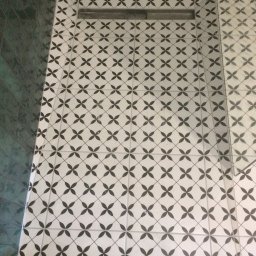 NiekraszBud - Białe płytki podłogowe w łazience z wzorem czarnych czterolistnych koniczynek i odpływem liniowym w rogu, kontrastujące z ciemnozielonymi płytkami ściennymi.