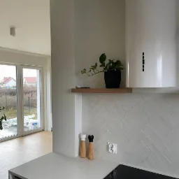 Nowoczesna kuchnia z białymi płytkami ułożonymi w jodełkę na ścianie, drewnianą półką z zieloną rośliną w czarnej doniczce, białym okapem, solniczką i pieprzniczką na blacie oraz widokiem...