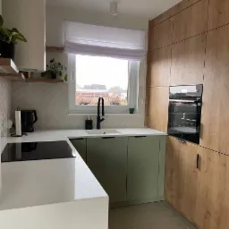 Nowoczesna kuchnia z zielonymi szafkami, drewnianą zabudową, białym blatem i czarną armaturą, wykończona z dbałością o detale.