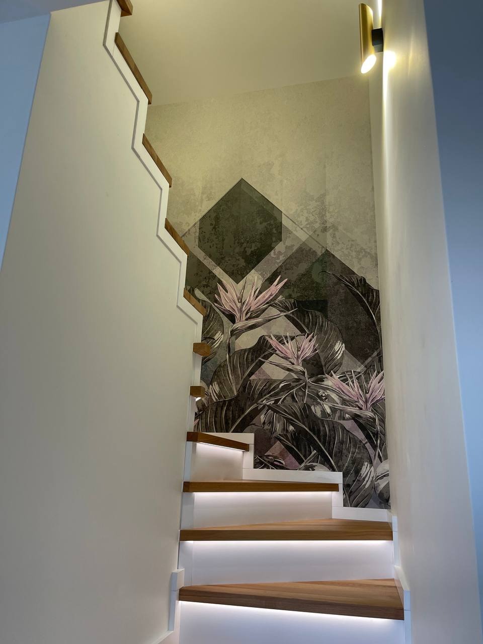 Schody z drewnianymi stopniami, podświetlone LED, z tapetą w motywy roślinne i geometryczne na ścianie.