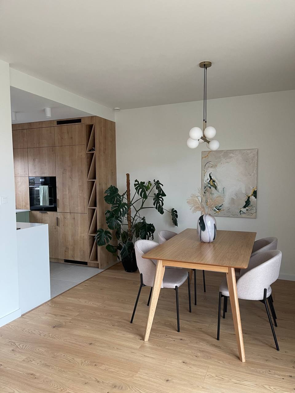 Jasne wnętrze jadalni z drewnianym stołem, czterema krzesłami z jasną tapicerką i nowoczesną lampą wiszącą. W tle zabudowa kuchenna z drewnianymi frontami i duża roślina doniczkowa.