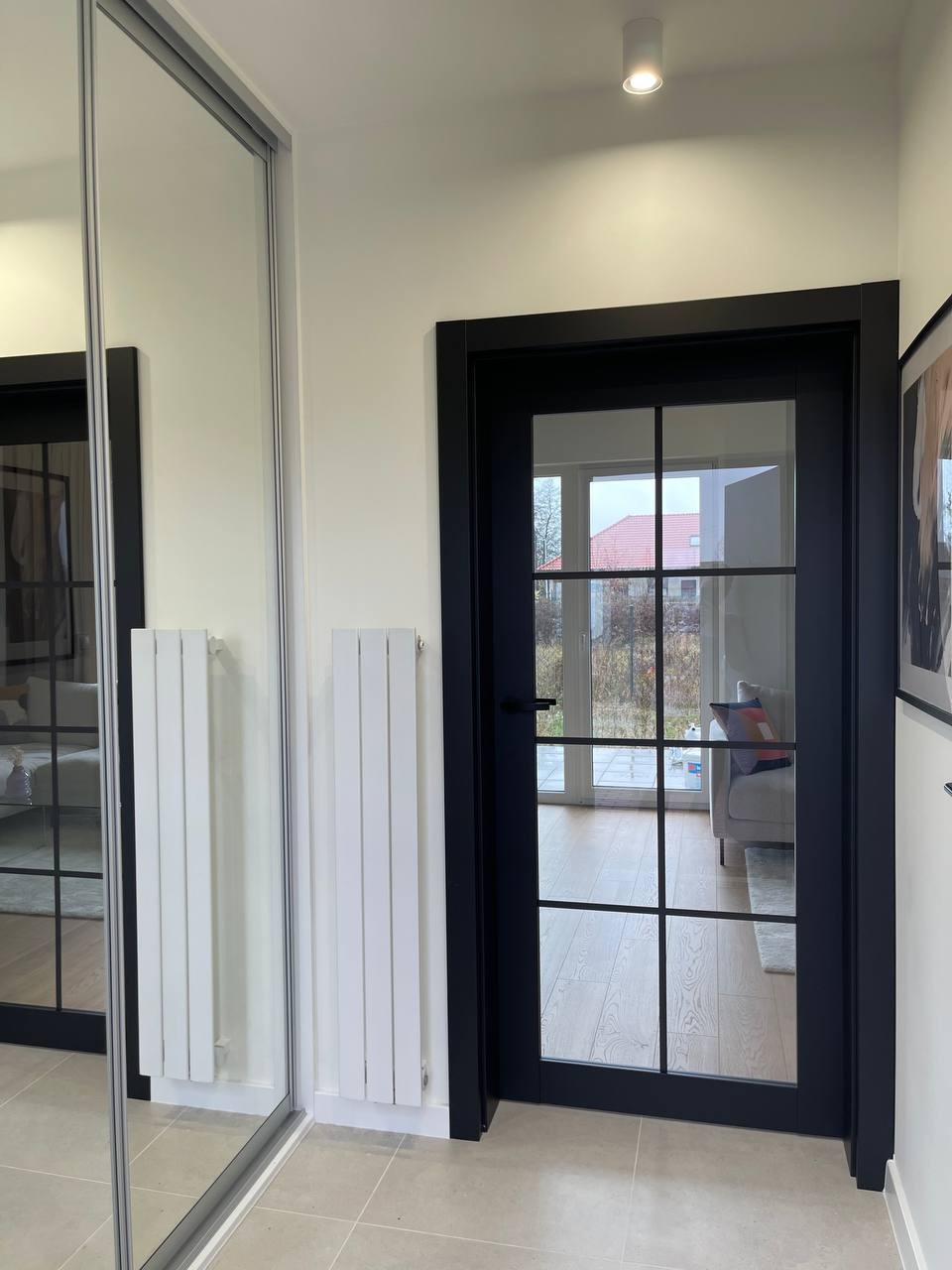 Nowoczesny przedpokój z przesuwną szafą z lustrem, białymi grzejnikami i czarnymi drzwiami przeszklonymi w stylu loft, widok na salon.