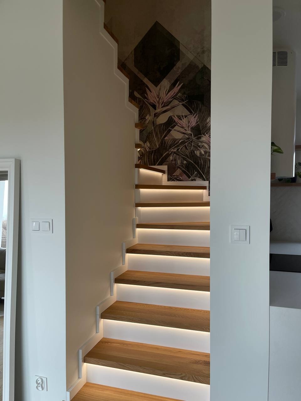 Schody drewniane z podświetleniem LED, widok z dołu, na ścianie tapeta z motywem tropikalnym, nowoczesne wykończenie.
