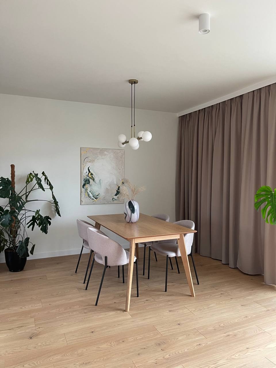 Jasne wnętrze jadalni z drewnianym stołem, krzesłami w odcieniach różu, abstrakcyjnym obrazem na ścianie i parkietem o naturalnym odcieniu drewna, oświetlone nowoczesną lampą wiszącą z kulistymi...