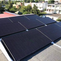 Słoneczny Doradca PL Sp. z o.o. - Cztery rzędy czarnych paneli fotowoltaicznych na płaskim dachu pokrytym papą, zamontowane na białej, metalowej konstrukcji nośnej, w tle budynki mieszkalne i drzewa w słoneczny dzień.