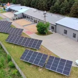 Widok z góry na instalację paneli słonecznych na trawie obok budynku przemysłowego i zaparkowany samochód z logo firmy.