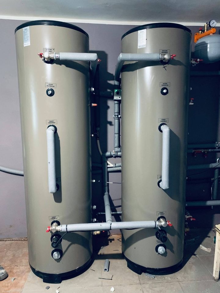 Dwa pionowe zbiorniki buforowe w kotłowni, połączone rurami z izolacją termiczną, widok z dołu, fragment buta w lewym dolnym rogu.