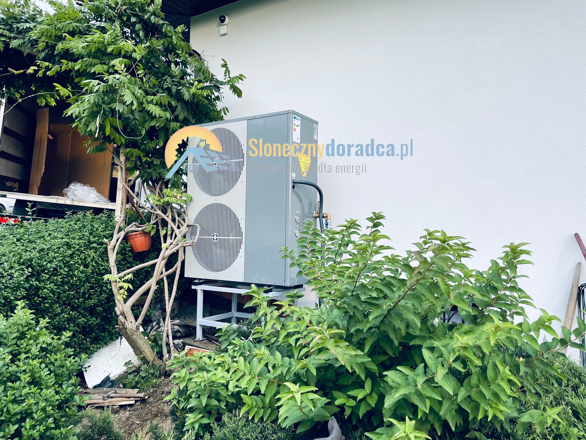 Szara pompa ciepła powietrze-woda zamontowana na metalowej konstrukcji przy białej ścianie budynku, częściowo zasłonięta przez zieloną roślinność i drzewo z doniczką, z widocznym logo firmy...