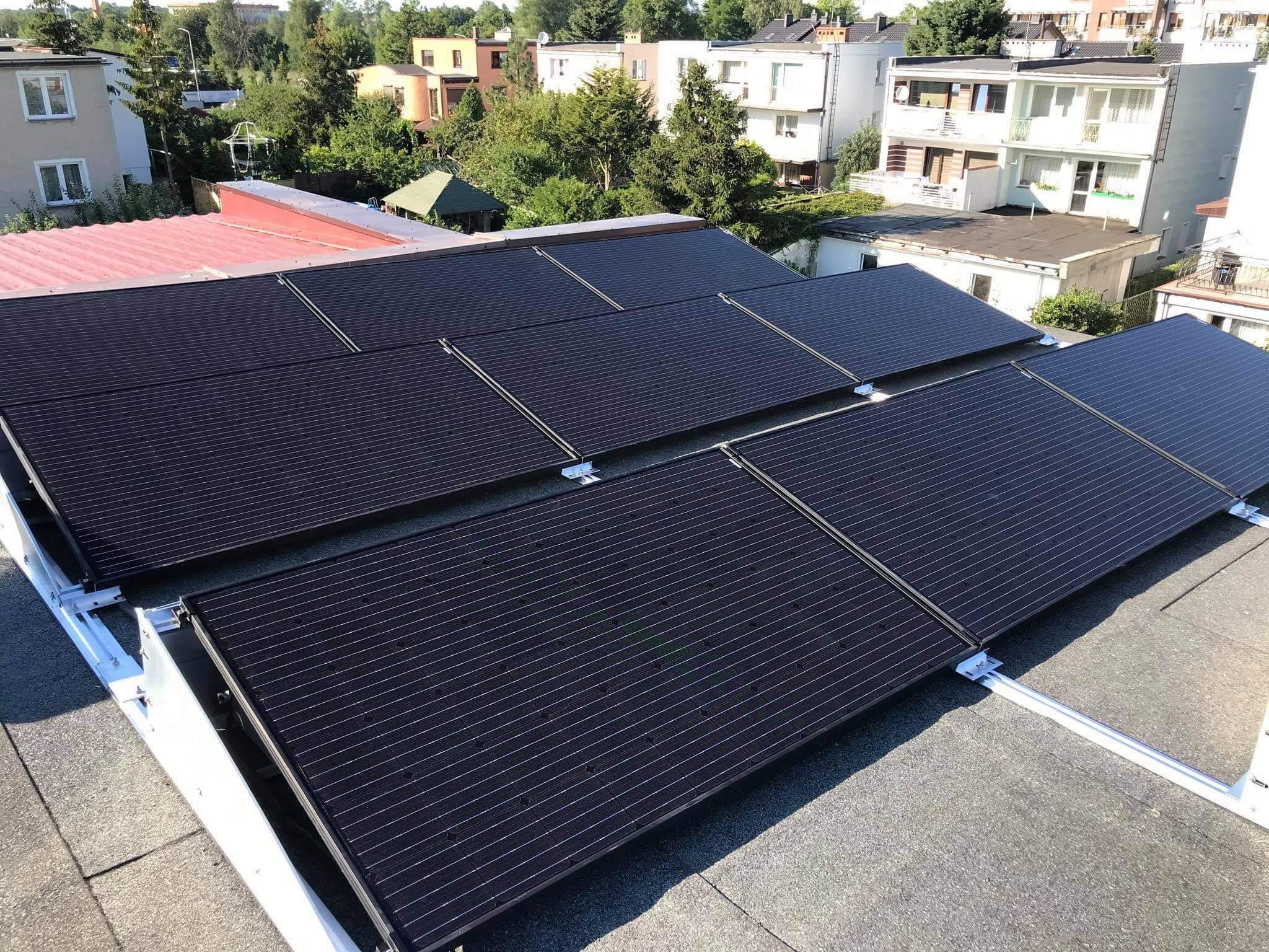 Cztery rzędy czarnych paneli fotowoltaicznych na płaskim dachu pokrytym papą, zamontowane na białej, metalowej konstrukcji nośnej, w tle budynki mieszkalne i drzewa w słoneczny dzień.