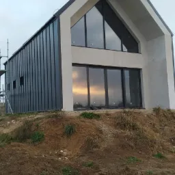 Nowoczesny dom szkieletowy w trakcie budowy, widoczne metalowe panele elewacyjne, duże okna z odbijającym się zachodem słońca, ziemny teren przed budynkiem i rusztowanie z lewej strony.