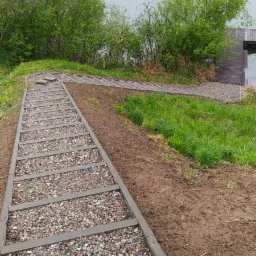 Schody terenowe z kamieniami prowadzące w dół zbocza w kierunku jeziora, z widoczną drewnianą platformą na brzegu.