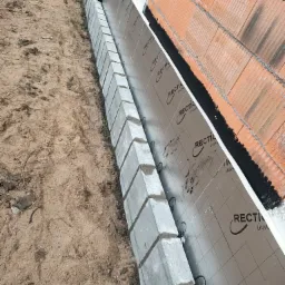 Wykop pod fundament z ułożonymi bloczkami betonowymi, izolacją przeciwwilgociową i ścianą z czerwonej cegły dziurawki w trakcie budowy.