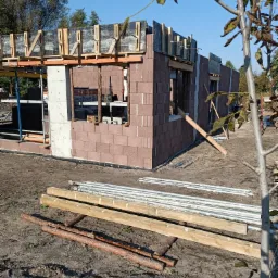 Wznoszenie ścian parterowego budynku z bloczków, widoczne szalunki na betonowe nadproża okienne i strop, w tle rusztowania i błękitne niebo.