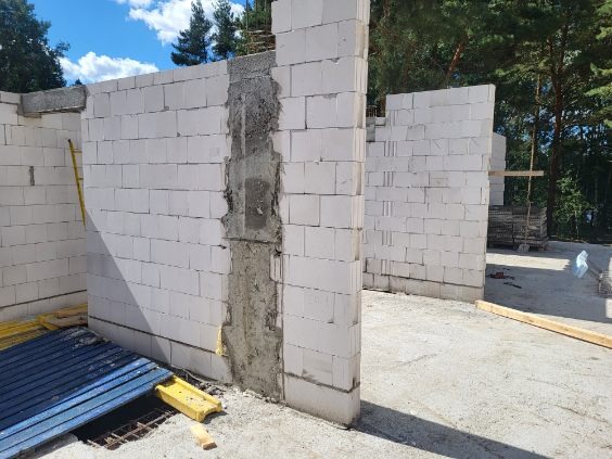 Wznoszenie ścian parteru z białych bloczków silikatowych na budowie domu jednorodzinnego, widoczne zbrojenie w narożniku, elementy szalunku i podkładki poziomujące.