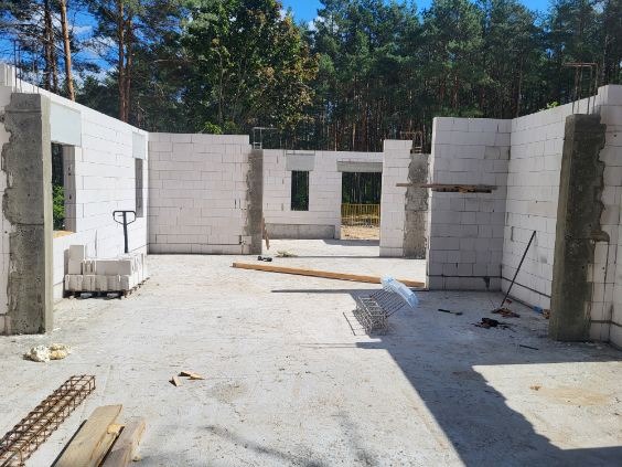 Wnętrze surowego domu w trakcie budowy z betonu komórkowego, widoczne ściany, słupy i otwory okienne, w tle las.