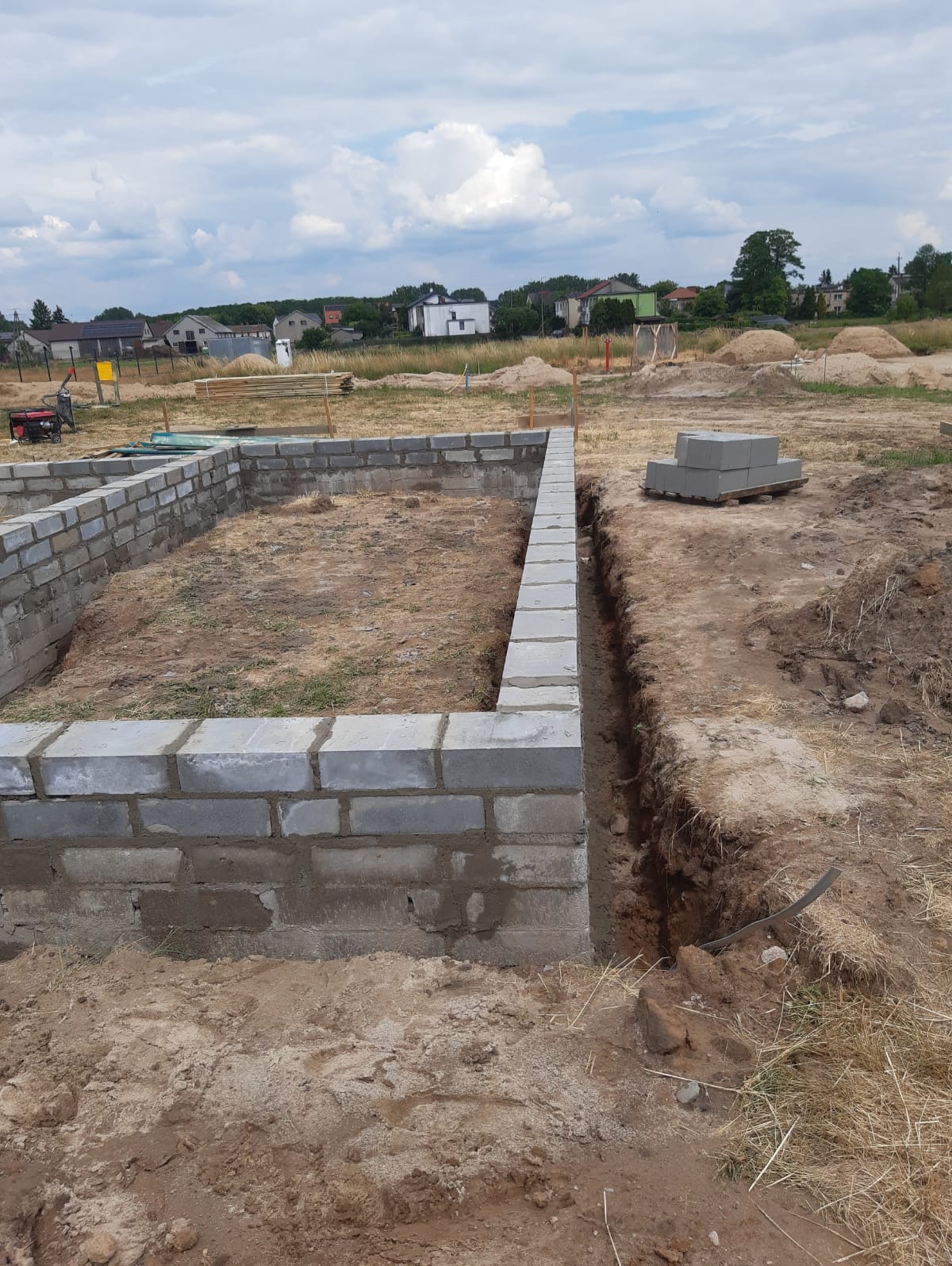 Wznoszenie fundamentów z bloczków betonowych na placu budowy, widoczny wykop i paleta z bloczkami w tle.