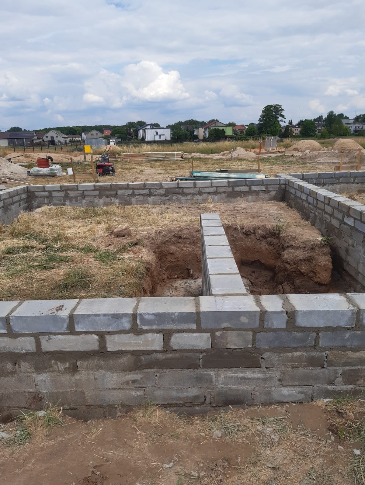 Wykopane fundamenty z częściowo wzniesionymi ścianami z szarych bloczków betonowych na placu budowy, widoczne narzędzia i materiały budowlane w tle.