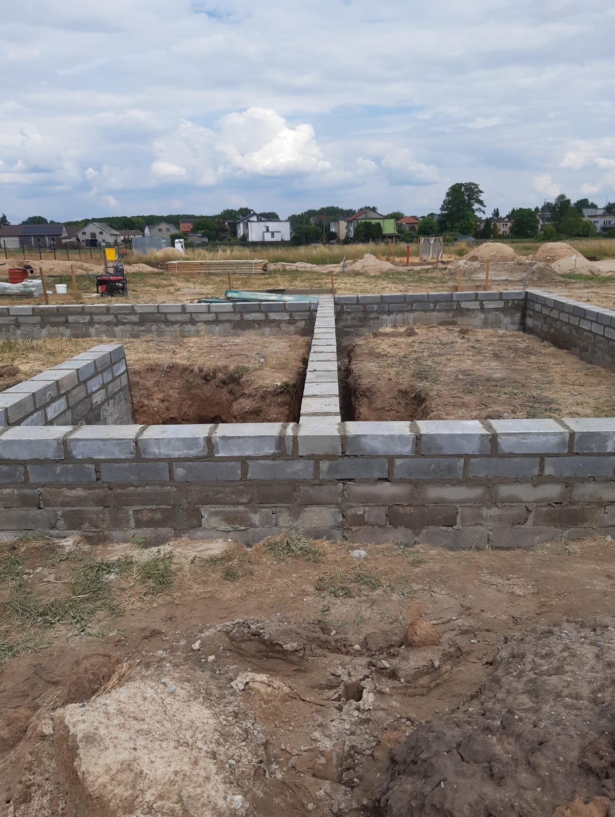 Wykop pod fundamenty z częściowo wymurowanymi ścianami z szarych bloczków betonowych, widoczne narzędzia i sprzęt budowlany w tle, krajobraz wiejski.