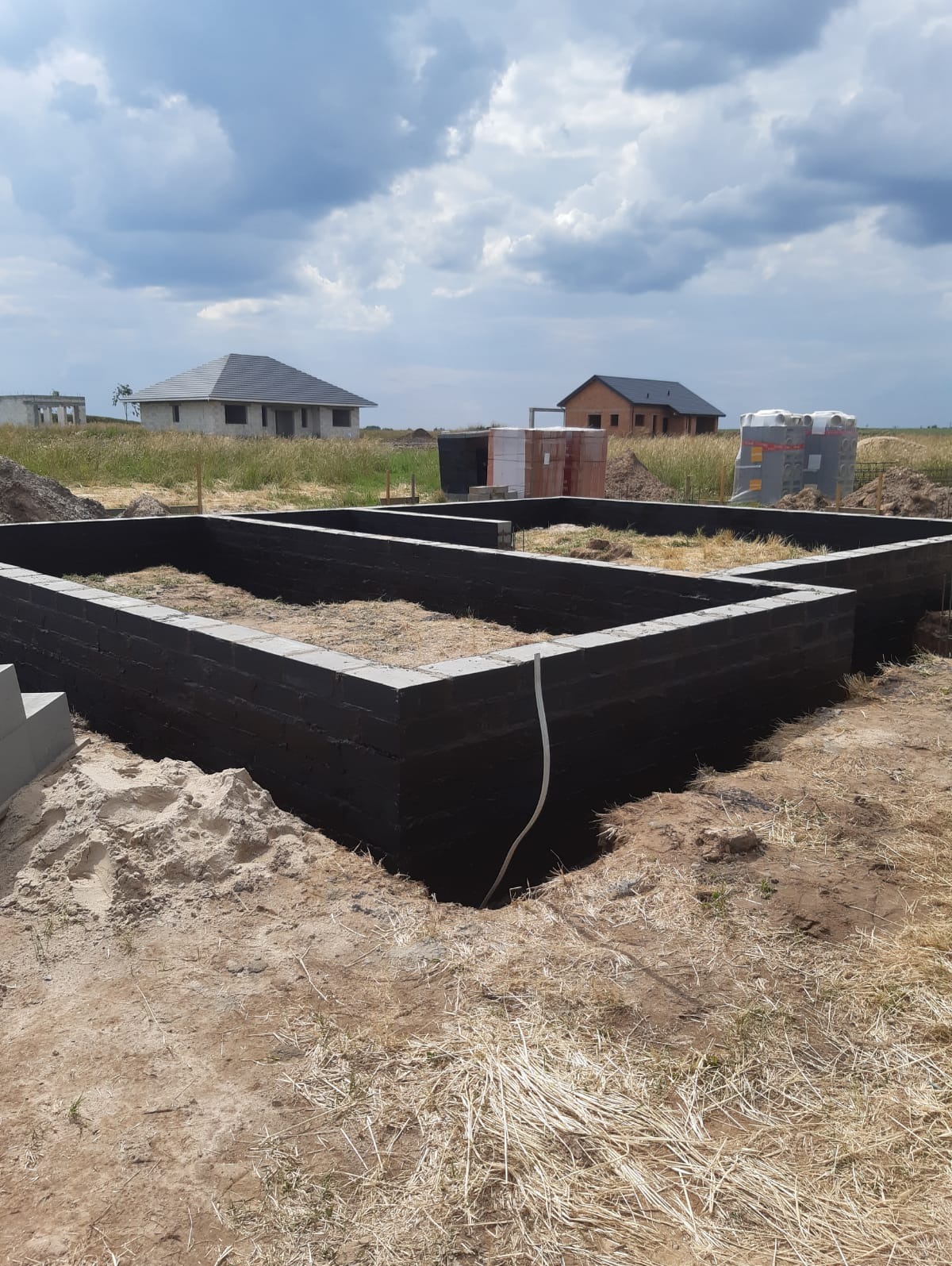 Zalane fundamenty z bloczków betonowych, zabezpieczone czarną izolacją, wypełnione słomą, na tle dwóch domów w stanie surowym i pochmurnego nieba.