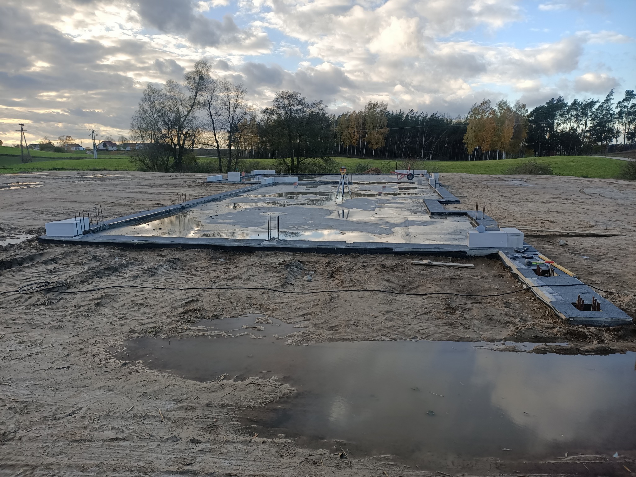 Zalane fundamenty domu w trakcie budowy, widoczne wystające pręty zbrojeniowe i białe bloczki fundamentowe, w tle zielony krajobraz i pochmurne niebo.