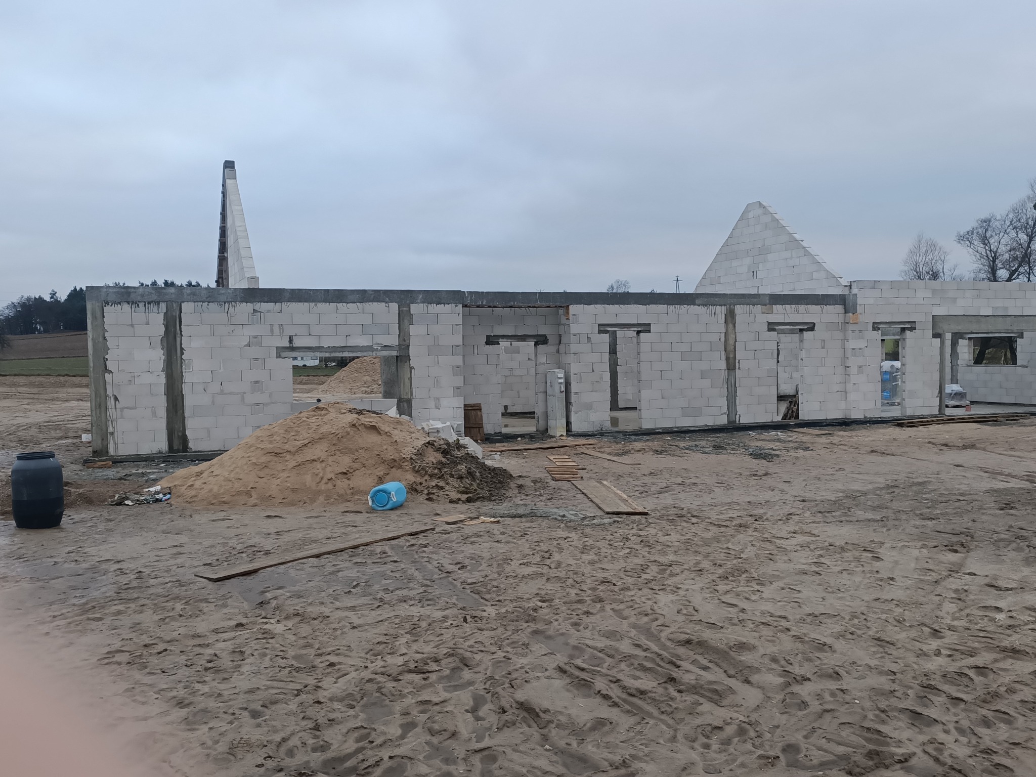 Wznoszone mury domu murowanego z betonu komórkowego, widoczny szczyt dachu w tle, hałda piasku i elementy wyposażenia budowy na nieutwardzonym terenie.