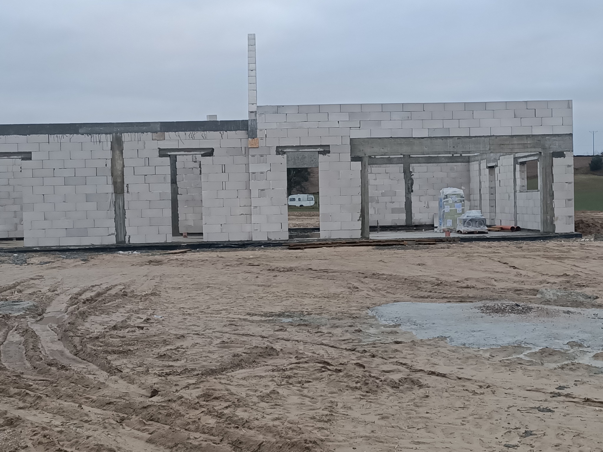 Surowy stan budowy domu z białych bloczków, widoczne otwory okienne i drzwiowe, z tyłu przyczepa kempingowa, ziemisty teren budowy z kałużami.