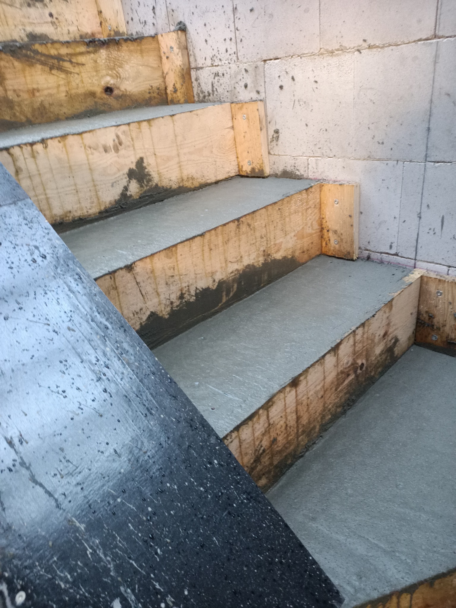 Wypełnianie betonem drewnianego szalunku schodów zewnętrznych, widoczne ślady betonu na deskach i ścianie z bloczków.