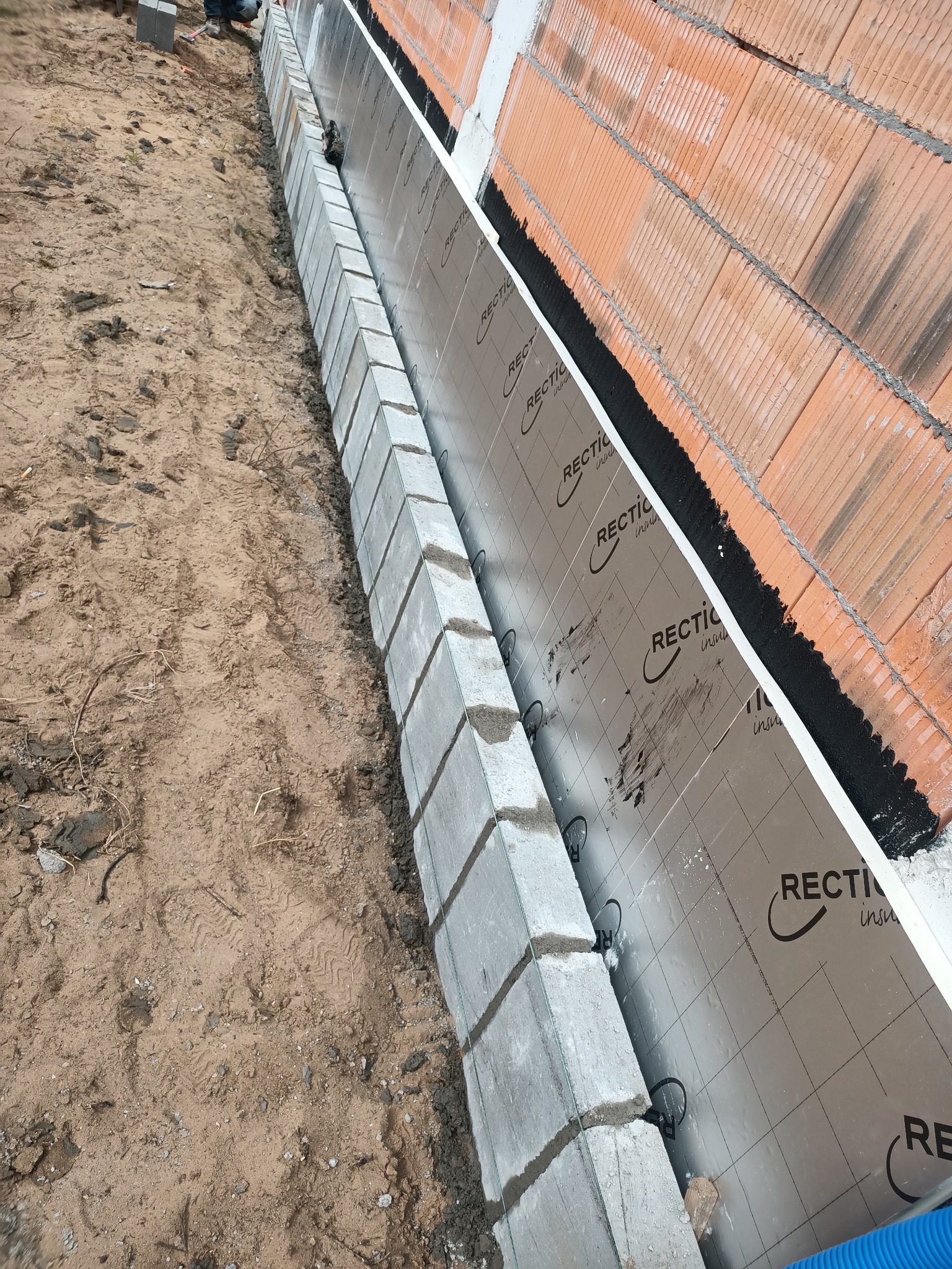 Wykop pod fundament z ułożonymi bloczkami betonowymi, izolacją przeciwwilgociową i ścianą z czerwonej cegły dziurawki w trakcie budowy.