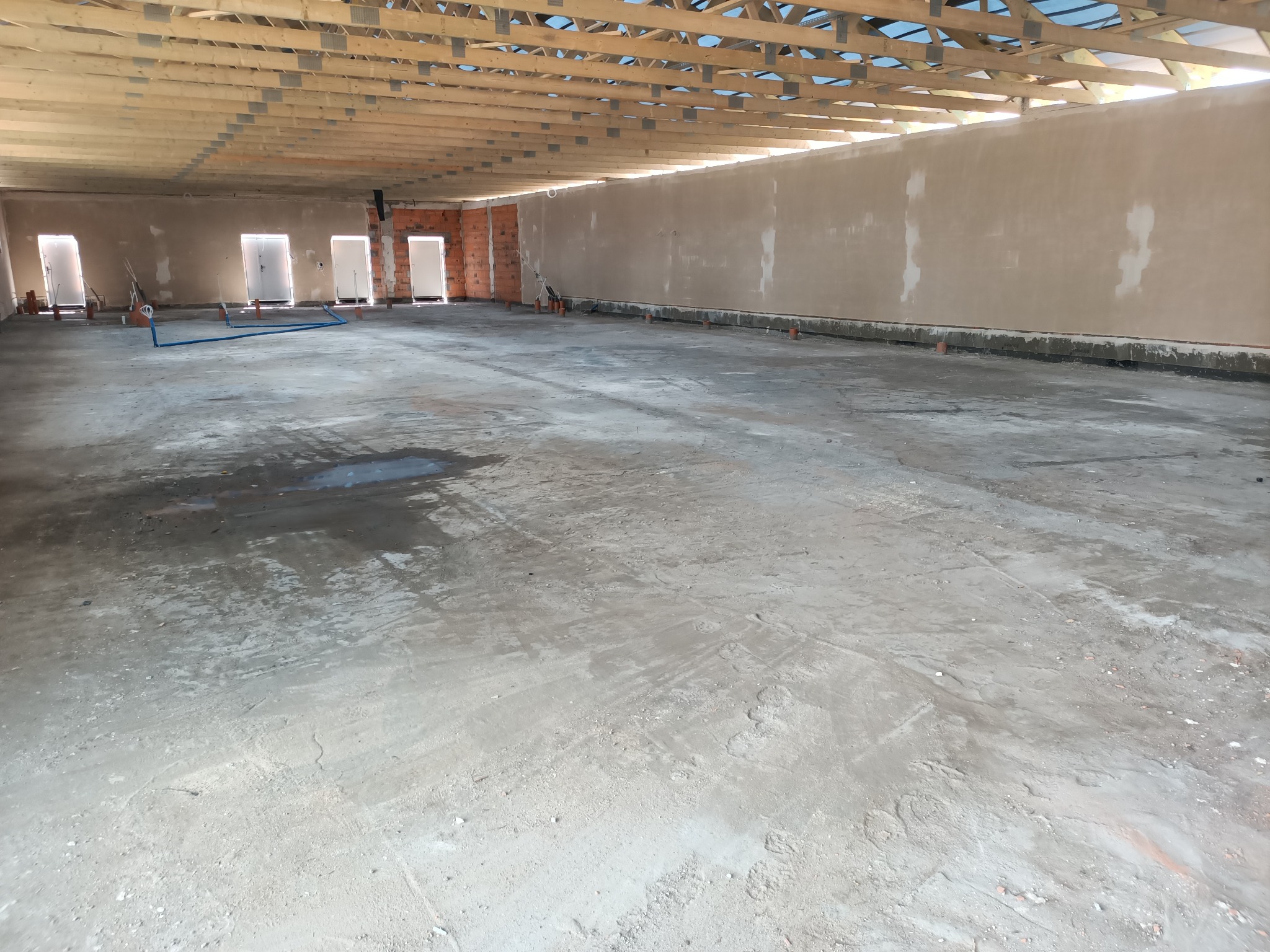 Wnętrze surowego budynku w trakcie budowy z widocznymi drewnianymi elementami konstrukcyjnymi dachu, otynkowanymi ścianami, betonową posadzką z kałużami i otworami drzwiowymi.